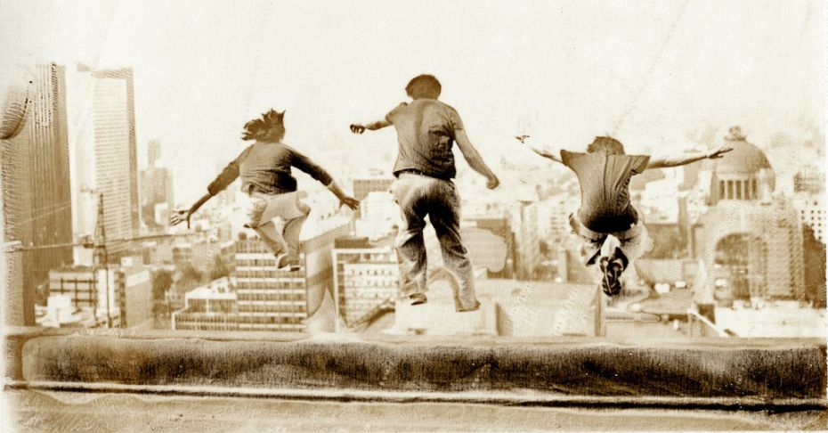 Jedna z fotografii Tomasza Gudzowatego przedstawiająca trzy sylwetki, którzy skaczą. w tle widoczna jest wysoka zabudowa dużego miasta